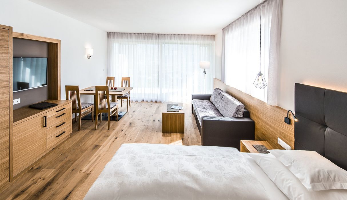 Hell eingerichtetes Hotelzimmer mit Bett, Sofa, Esstisch und großem Fenster mit durchsichtigen Vorhängen