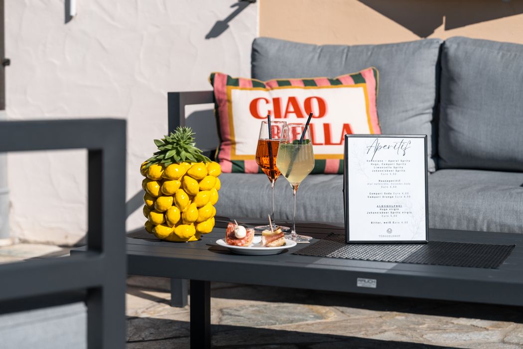 Sommerliche Terrasse mit grauem Sofa, Kissen mit Aufschrift 'Ciao Bella', gelber Dekopflanze, Getränken und Aperitifkarte auf schwarzem Tisch.