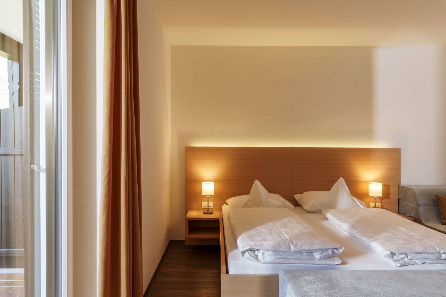 Hell eingerichtetes Schlafzimmer mit Doppelbett, zwei Nachttischen mit Lampen und großem Fenster mit Vorhang.