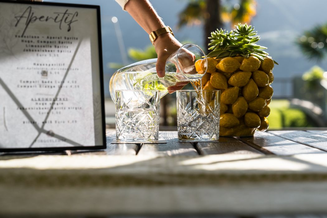 Hand gießt Wasser aus Karaffe in zwei Gläser auf Holztisch mit Dekoration und Aperitif-Menü
