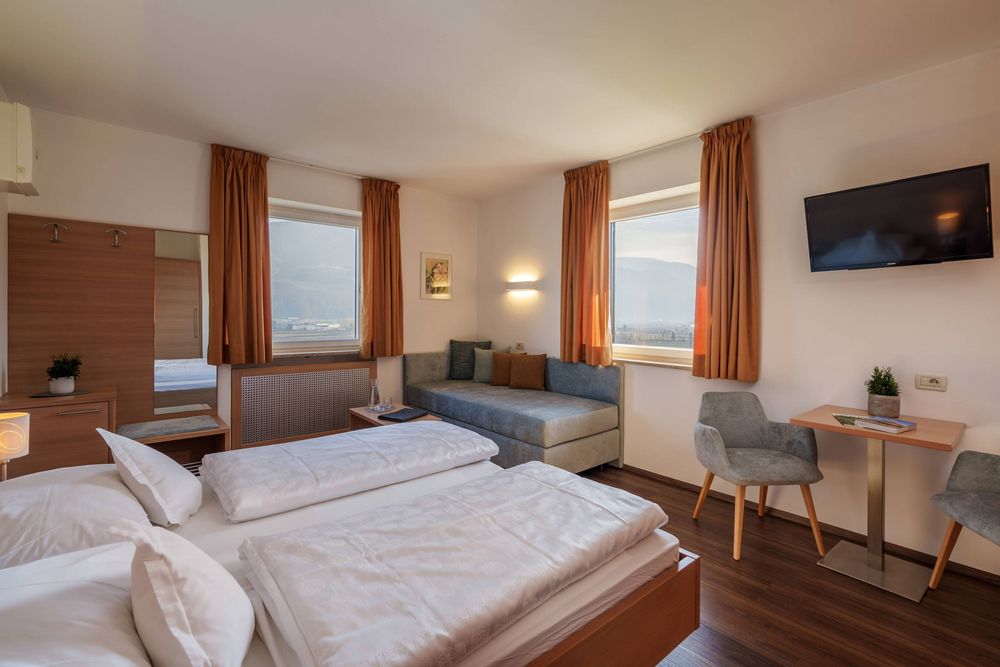 Cozy hotel room with double bed, sofa, two windows with orange curtains, small table with chairs, and wall-mounted TV.