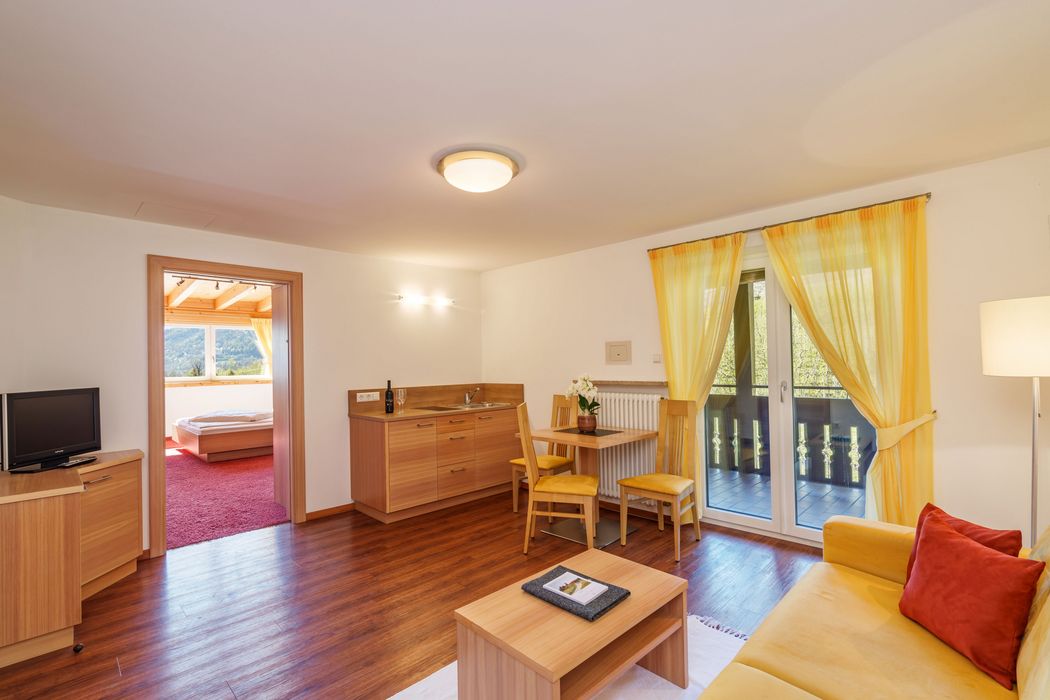Helles Wohnzimmer mit gelbem Sofa, kleinem Holztisch, Esstisch mit Stühlen, Küchenzeile und Zugang zu Balkon und Schlafzimmer.