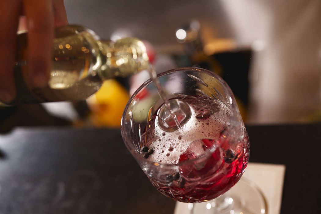 Nahaufnahme eines Glases mit rotem Getränk, Eiswürfeln und Beeren, in das Flüssigkeit eingeschenkt wird.