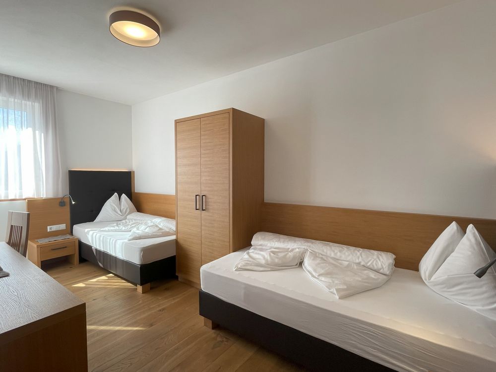 Camera d'albergo con due letti singoli, armadio in legno, pavimento in legno e finestra con tende bianche.