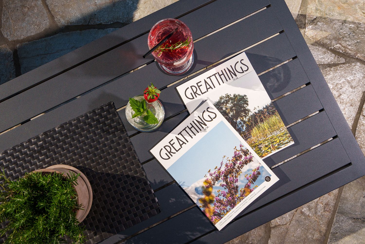 Zwei Magazine mit der Aufschrift 'Great Things' auf einem Gartentisch neben zwei Getränken und einem kleinen Pflanzenkübel.