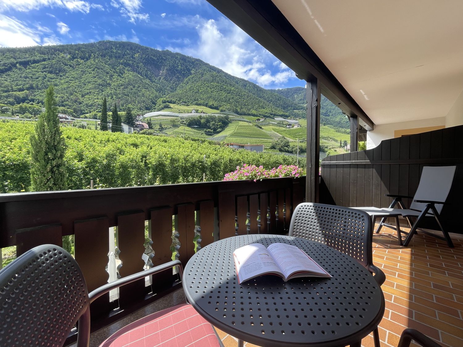 Balkon mit Tisch, Stühlen und einem offenen Buch, Blick auf grüne Hügel und blauen Himmel