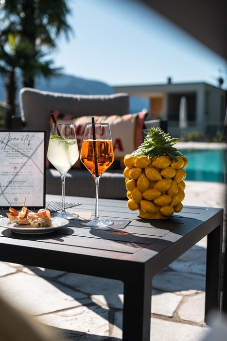 Sommerliche Terrasse mit zwei gefüllten Weingläsern, einer Ananas-Dekoration, kleinen Häppchen auf einem Teller und einem Aperitif-Menü, im Hintergrund Pool und Palmen.