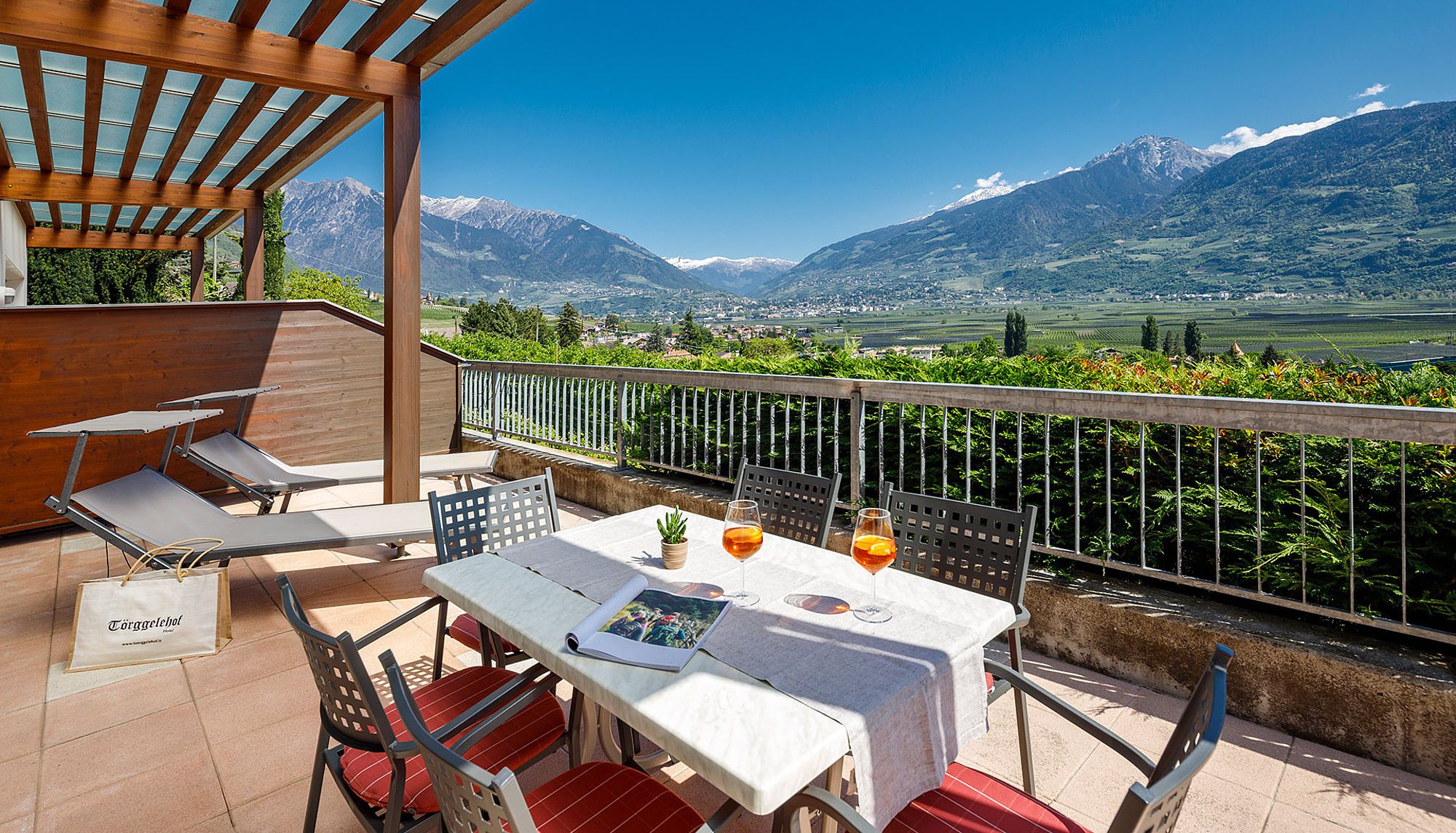 Terrasse mit Tisch, zwei Aperol-Gläsern, Liegestühlen und Bergblick unter blauem Himmel