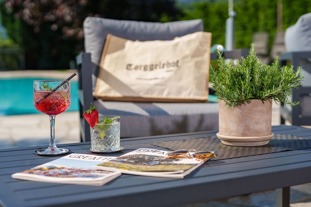 Getränke, Magazin und Pflanze auf Gartentisch vor gemütlichem Sessel und Pool