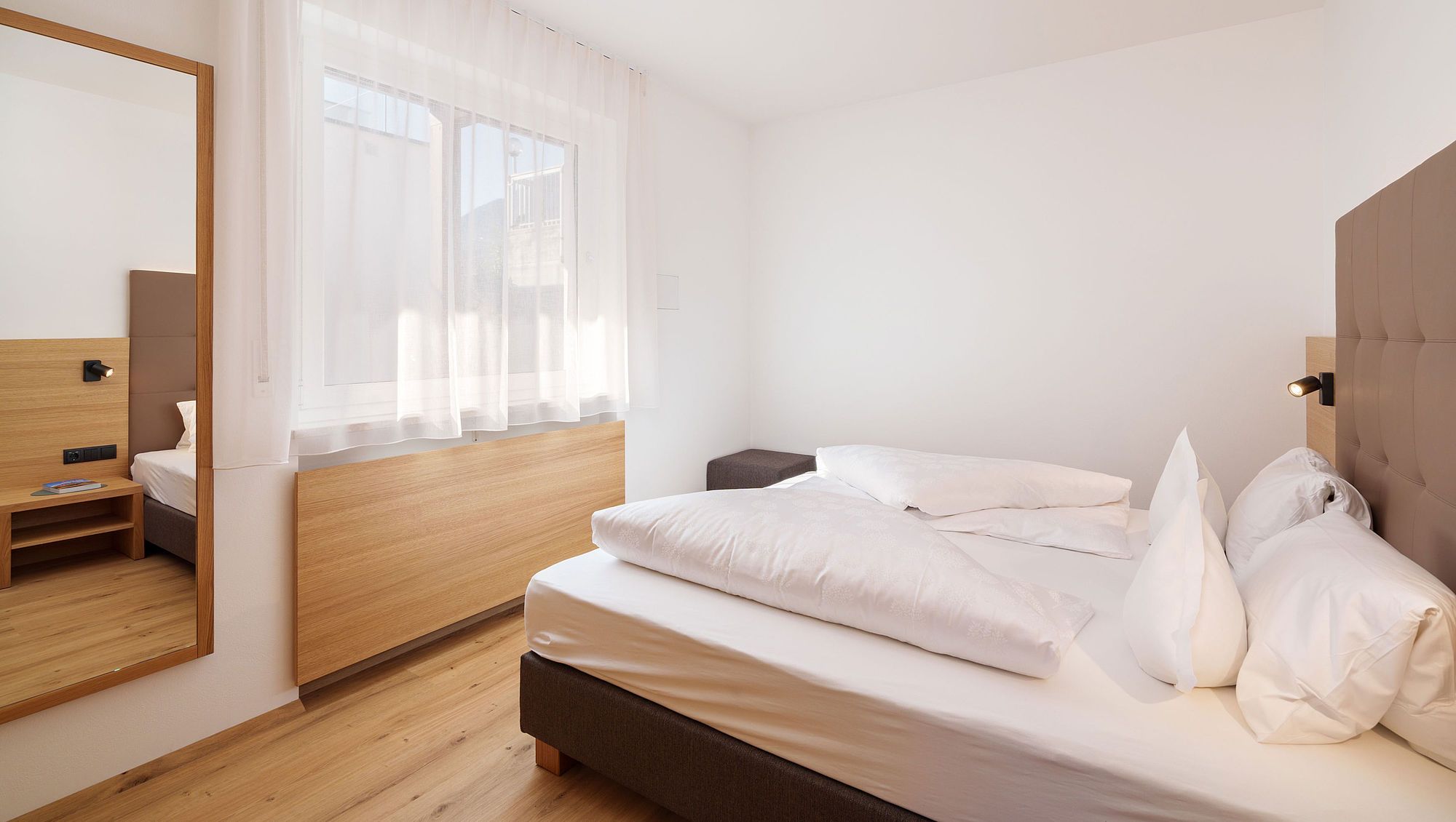 Helles Schlafzimmer mit Doppelbett, weißen Bettwäsche, großem Spiegel und Fenster mit Vorhängen.