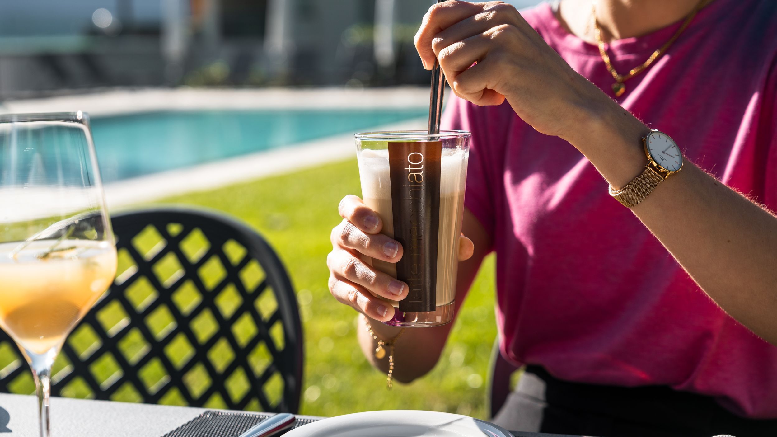 Person rührt Getränk in Glas mit Aufschrift 'latte macchiato' an einem Tisch im Freien bei Sonnenschein