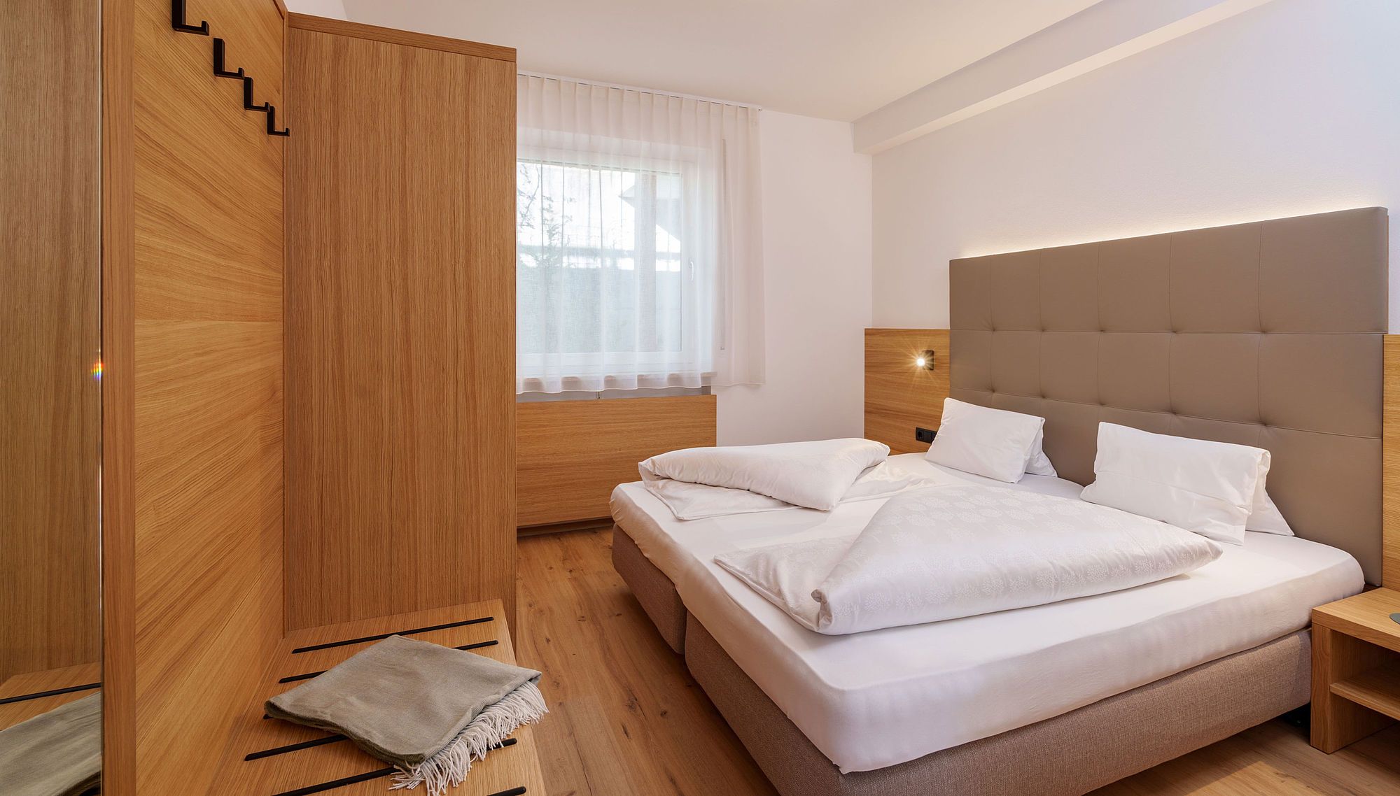 Modernes Schlafzimmer mit großem Bett, Holzmöbeln und einem Fenster mit weißem Vorhang.