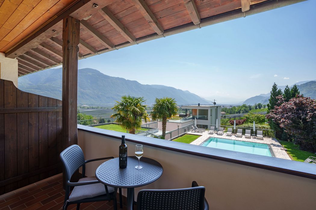 Balkon mit Tisch, zwei Stühlen, Weinflasche und Glas, Blick auf Pool, Palmen, Wiese und Berge bei klarem Himmel