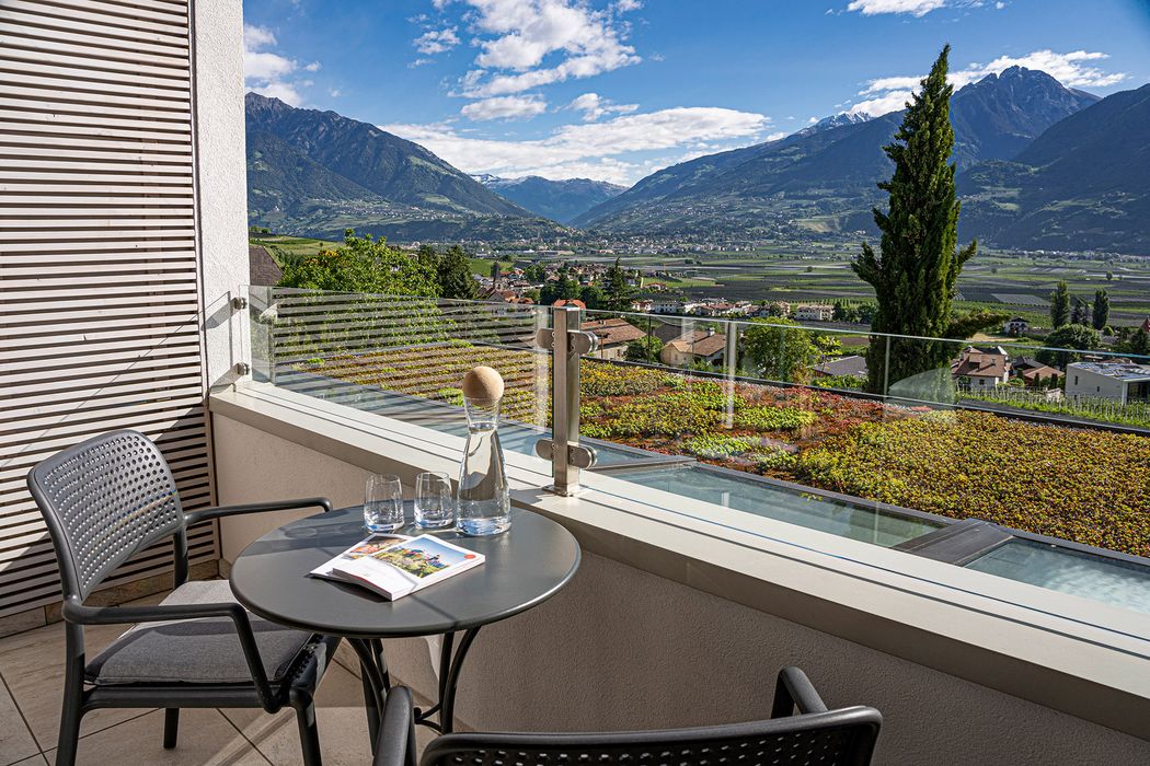 Balkon mit Tisch und zwei Stühlen, Blick auf ein Tal mit Häusern und Bergen im Hintergrund bei klarem Himmel