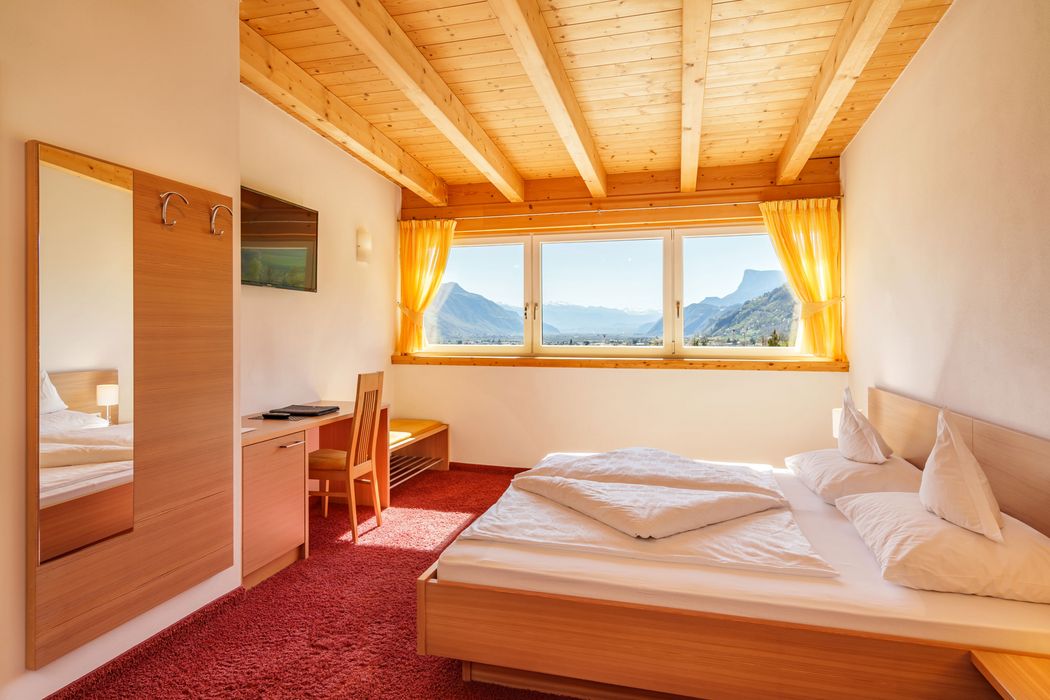 Helles Hotelzimmer mit Holzdecke, großem Fenster mit Bergblick, Doppelbett und rotem Teppich