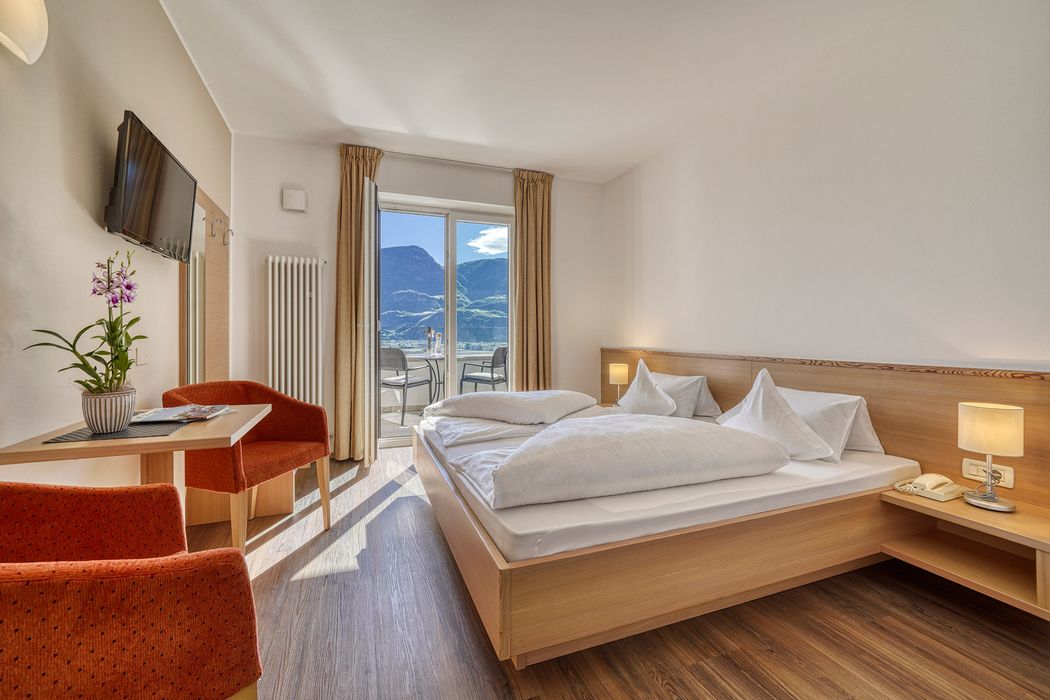 Helles Hotelzimmer mit Doppelbett, zwei roten Sesseln, Tisch mit Blume, TV und Balkon mit Bergblick