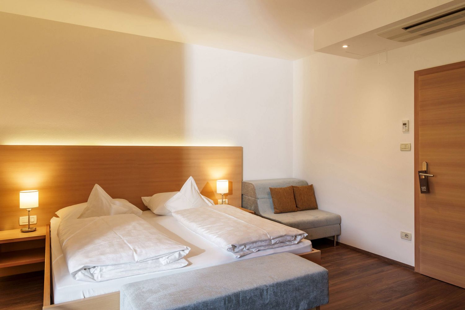 Helles Hotelzimmer mit Doppelbett, zwei Lampen, einer grauen Sitzbank und einem kleinen Sofa mit braunen Kissen.