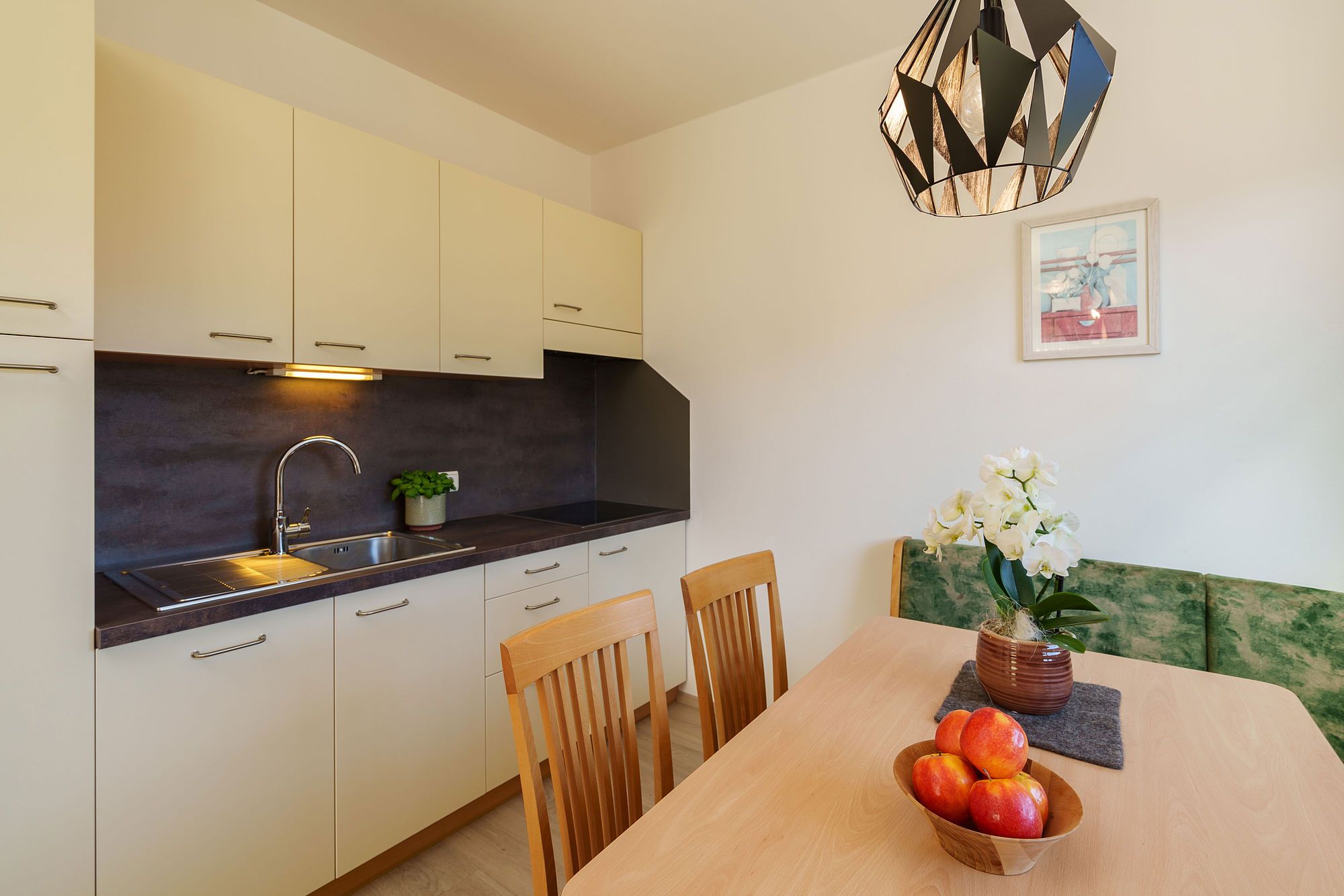 Moderne kleine Küche mit hellem Holz, weißen Schränken, grünem Sitzbank und Hängelampe, Tisch mit Äpfeln und weißer Blume.