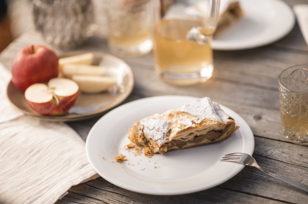 Ein Stück Apfelstrudel auf weißem Teller mit einer Gabel, im Hintergrund ein Teller mit frischen Apfelstücken und Getränke auf Holztisch.