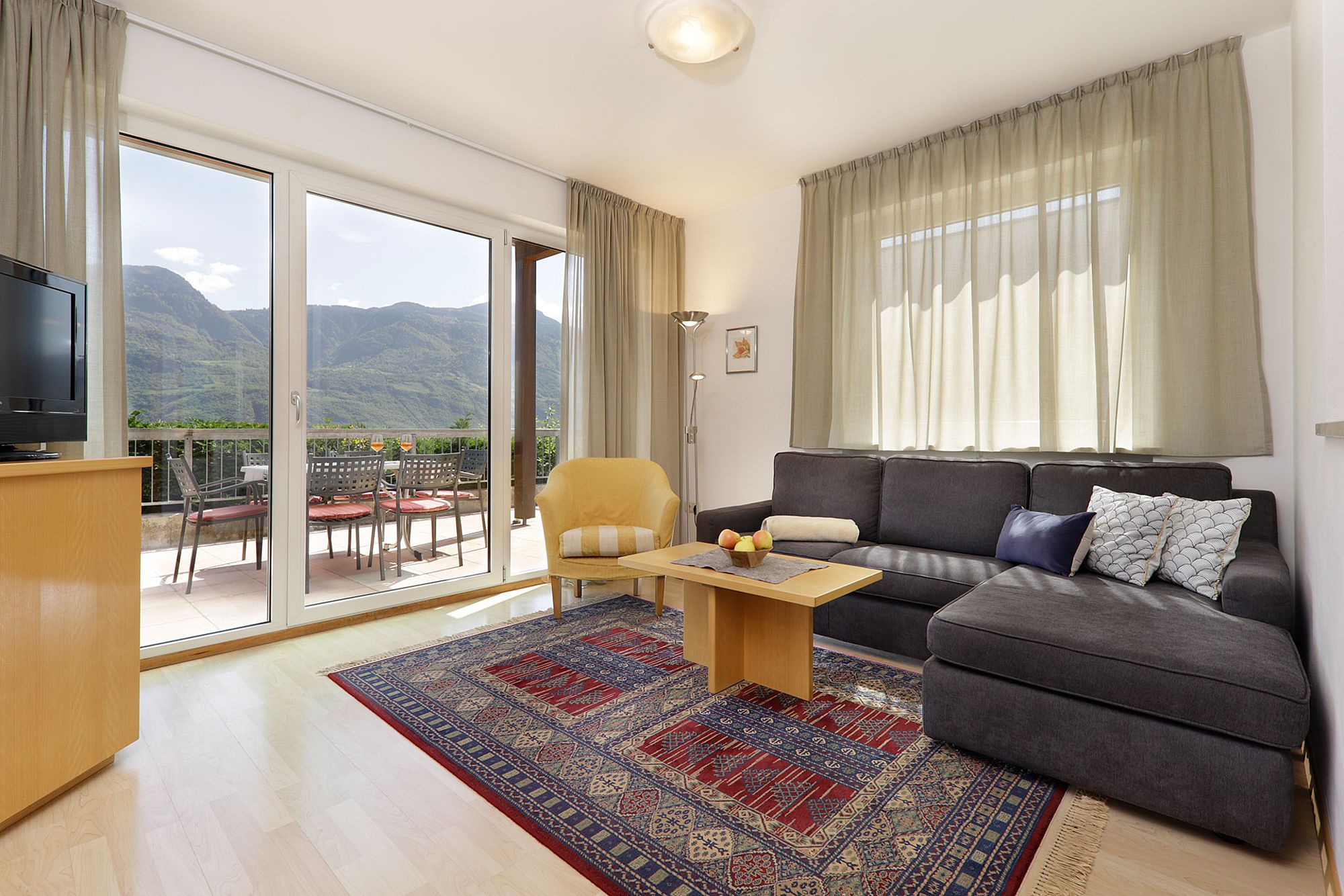 Helles Wohnzimmer mit grauer Eckcouch, gelbem Sessel, großem Fenster mit Blick auf Terrasse und Berge, Teppich mit orientalischem Muster, Holzfußboden und Fernseher.