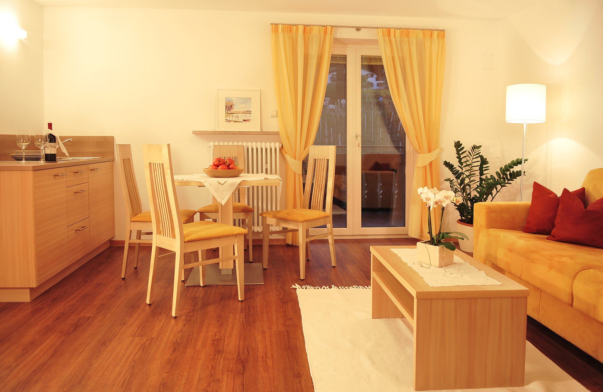 Gemütlicher Wohn- und Essbereich mit Holzmöbeln, gelben Vorhängen und Sofa, Tisch mit Blumen und Obstschale, Stehlampe und Pflanze.
