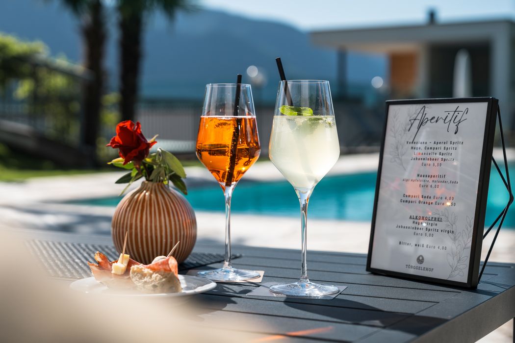 Zwei Aperitif-Gläser mit Strohhalmen, rote Rose in Vase, Smallplate mit Snacks und Aperitif-Menü am Tisch neben Pool im Freien