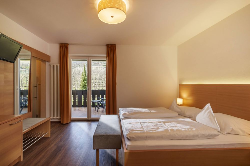Bright bedroom with wooden furniture, a double bed, a grey bench, floor-to-ceiling window with orange curtains, and a balcony with outdoor chairs.