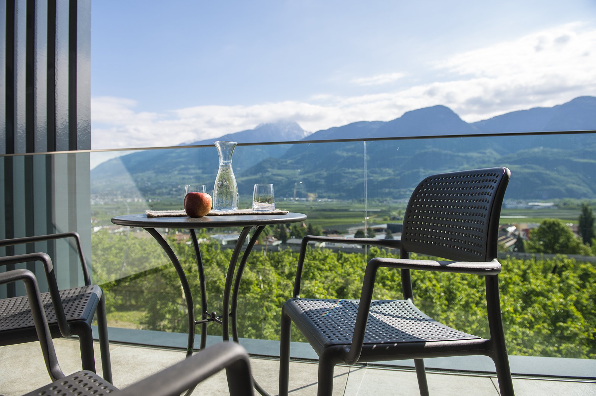 Balkon mit Tisch und Stühlen, darauf eine Karaffe, ein Glas und ein Apfel, mit Blick auf grüne Landschaft und Berge