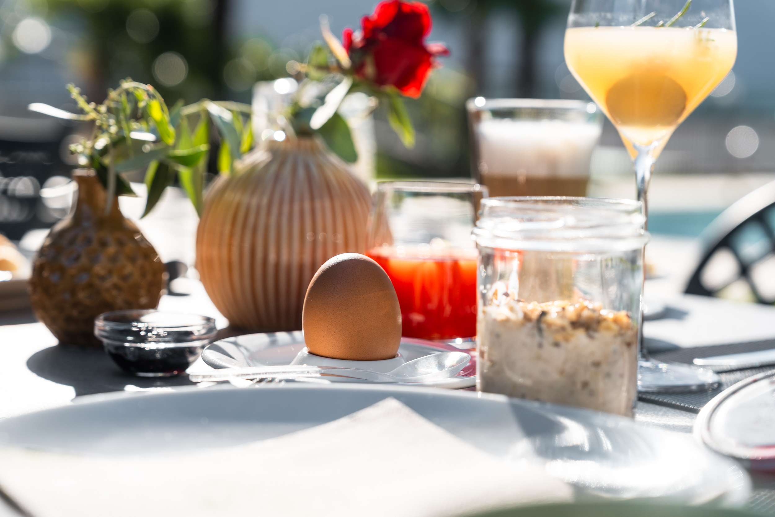 Frühstückstisch mit Ei, Müsli, Getränken und Vasen mit Blumen bei Sonnenlicht