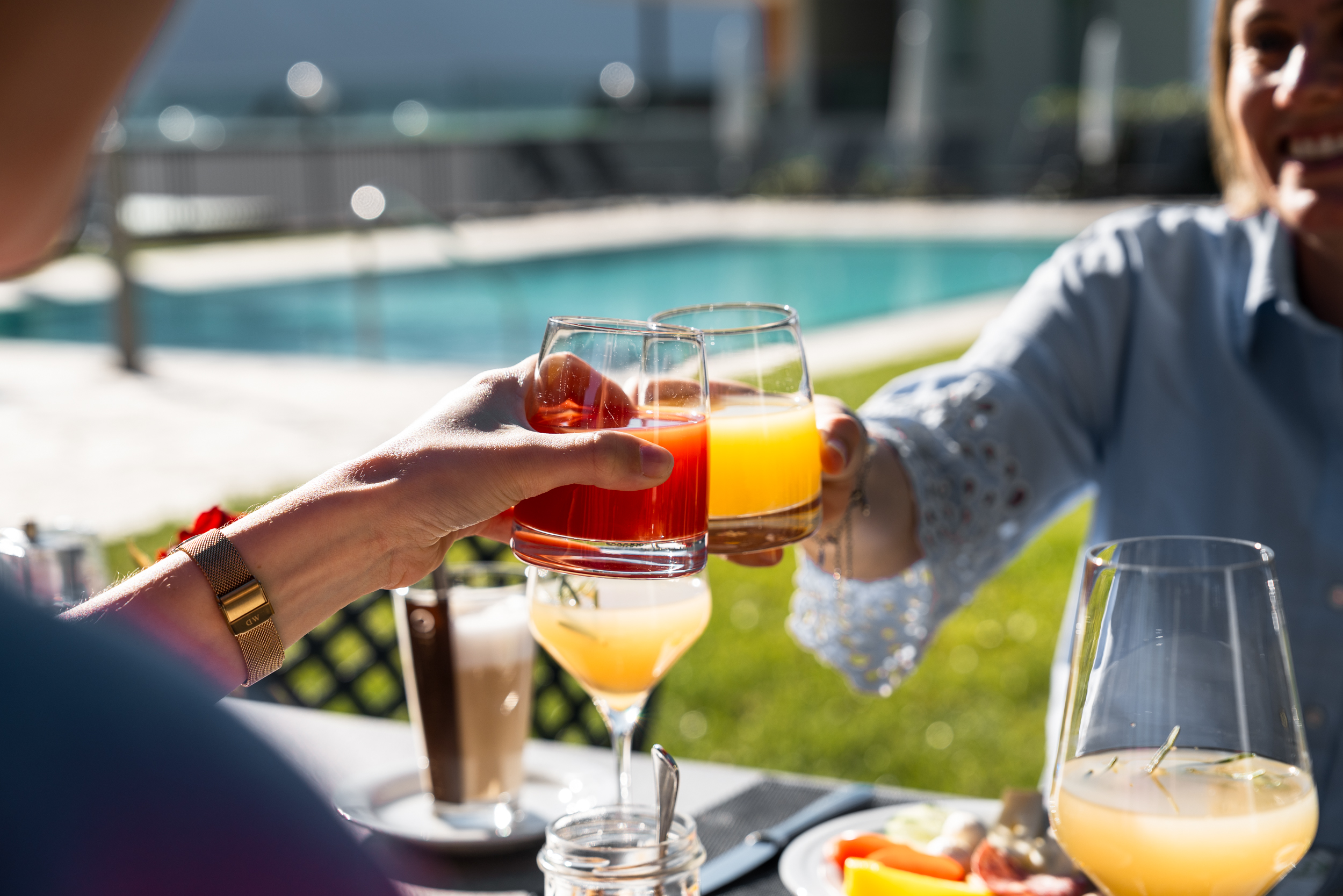 Zwei Personen stoßen mit bunten Getränken an einem Tisch im Freien bei Sonne an, im Hintergrund ein Pool.