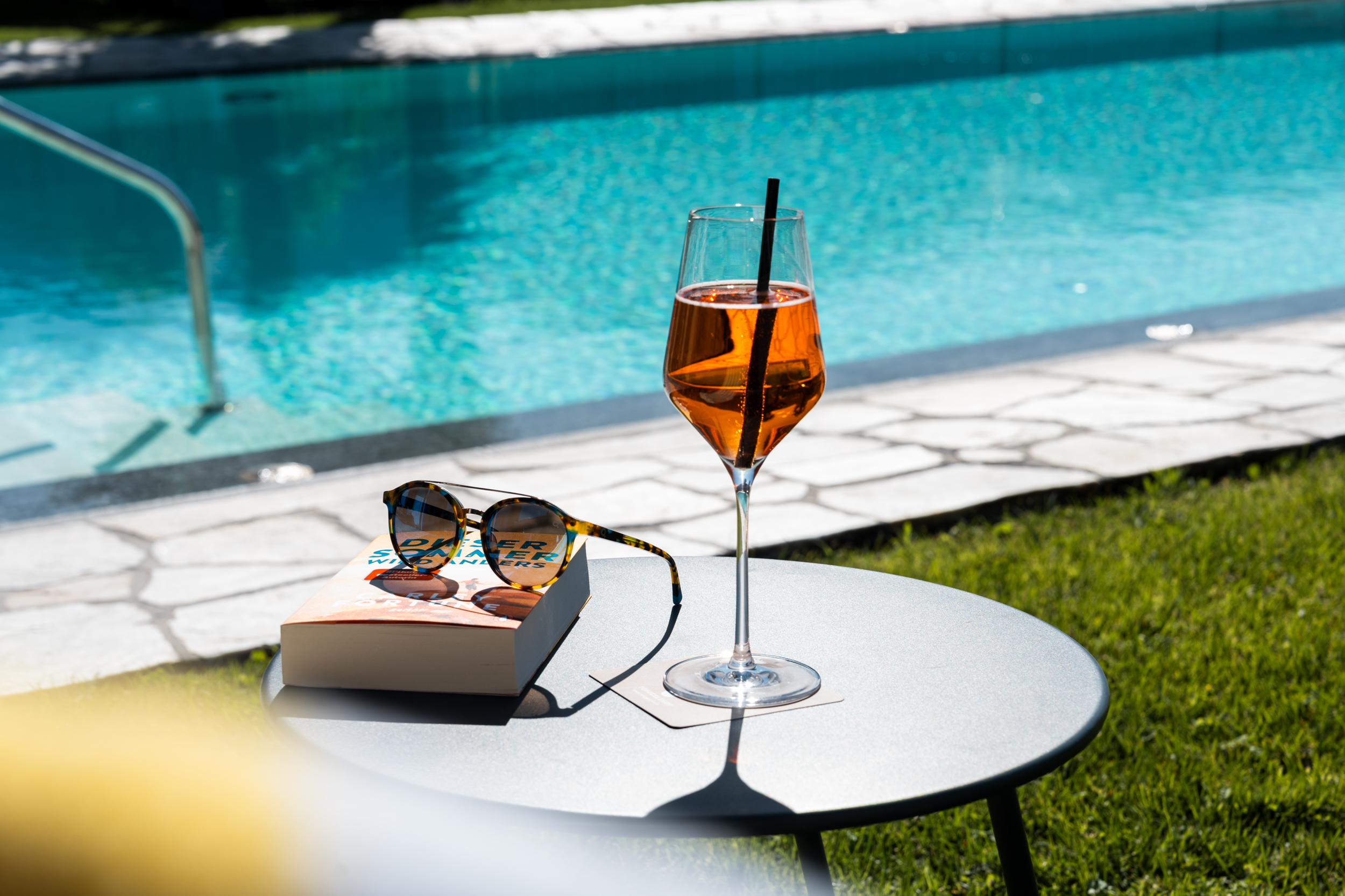 Cocktailglas mit orangem Getränk und Sonnenbrille auf Buch neben einem Pool