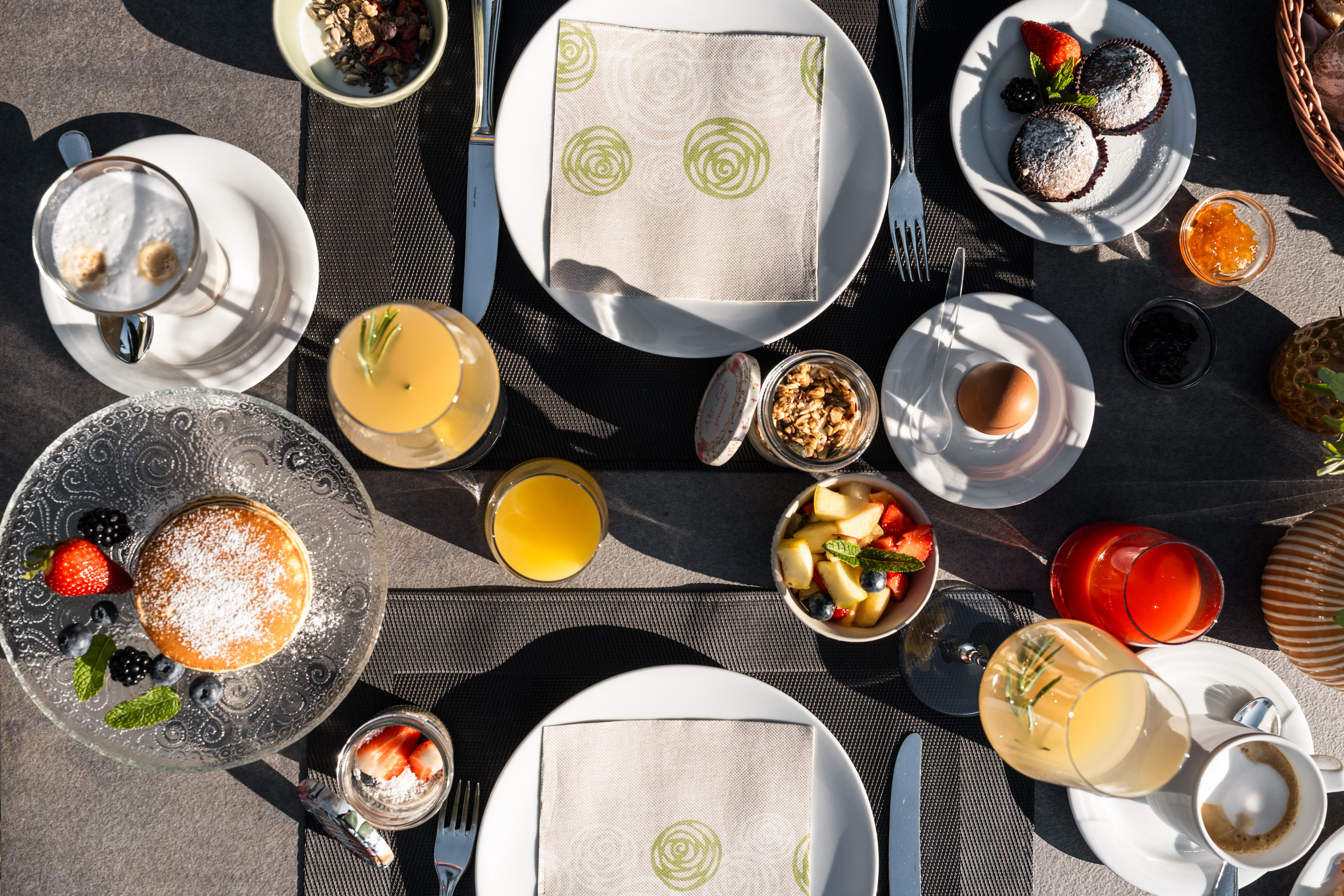 Frühstückstisch mit Pancakes, Muffins, Obstsalat, gekochtem Ei, Müsli, Kaffee und Säften, gedeckt mit Tellern und Besteck