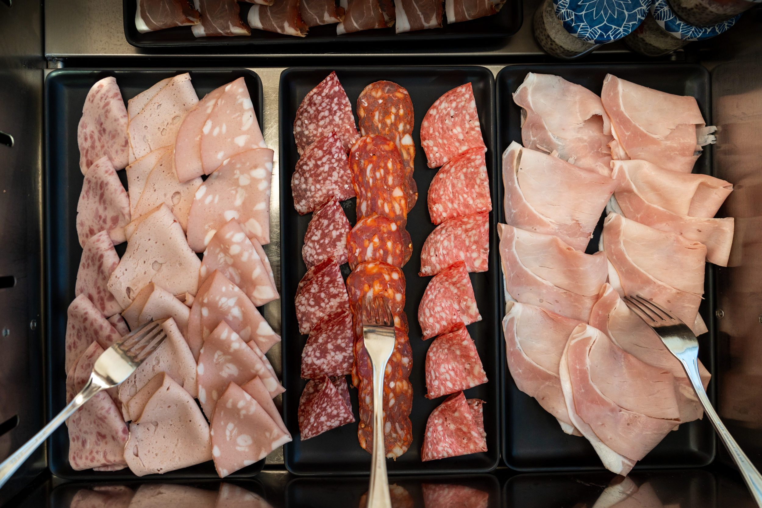 Drei schwarze Teller mit verschiedenen aufgeschnittenen Wurst- und Schinkensorten, Besteck liegt auf den Tellern.